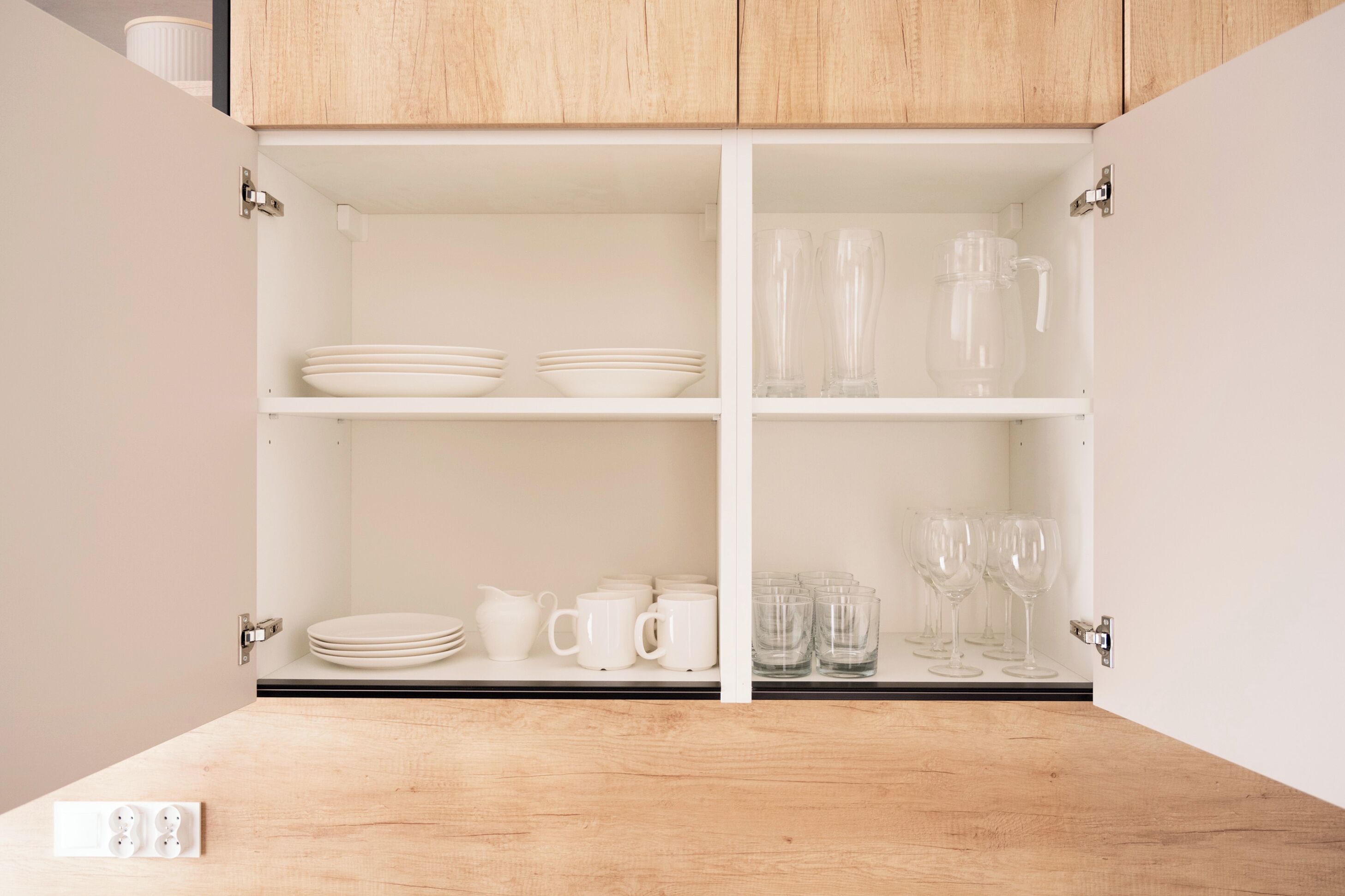 A kitchen with wooden cabinets, white shelves, and various kitchenware including plates, bowls, mugs, and glasses.