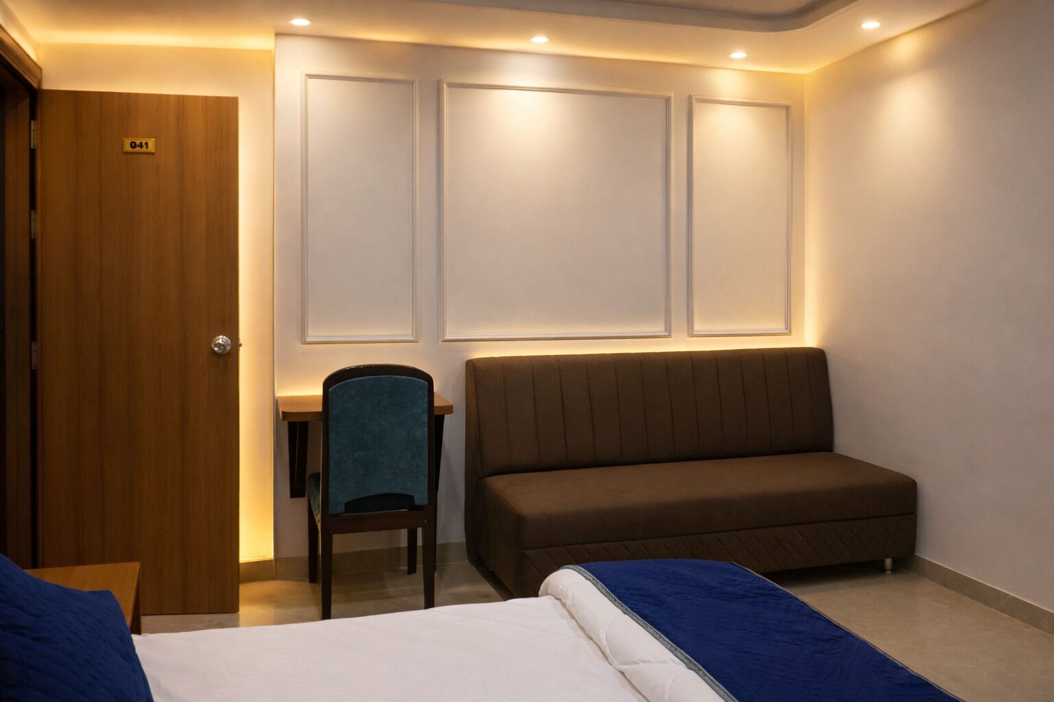 Une chambre d’hôtel avec une porte en bois, un bureau, une chaise bleue, un canapé marron et deux lits.