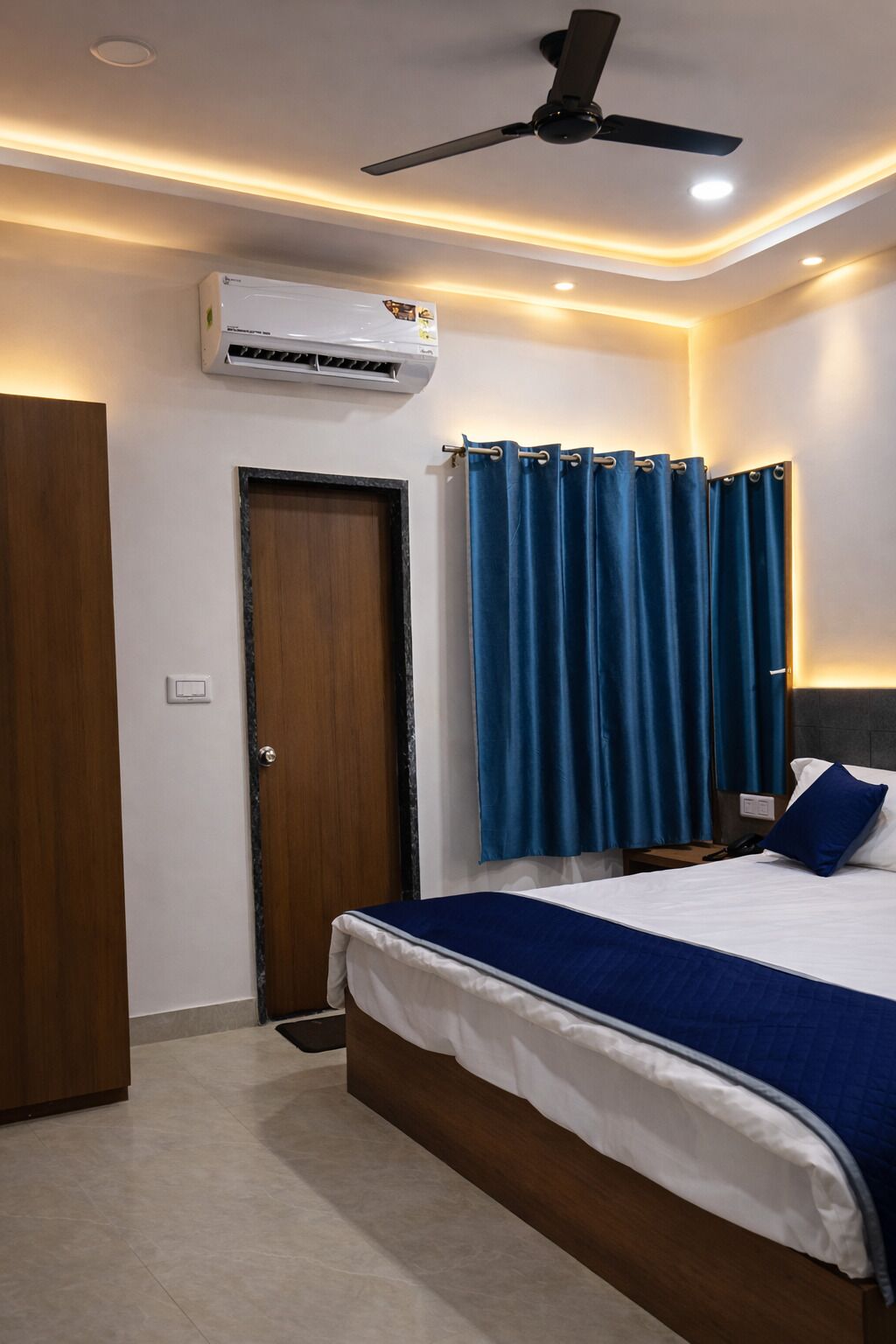 A bedroom with a bed, a ceiling fan, an air conditioning unit, a door, and blue curtains.