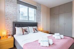 Un lit bien fait, recouvert d’une couverture rose et agrémenté de deux serviettes portant la mention « localhost », un mur décoré d’un motif en briques, une grande armoire et une fenêtre donnant sur des immeubles.