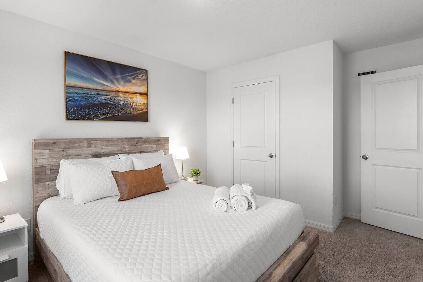Une chambre d’hôtel avec un lit doté d’une tête de lit en bois, du linge de lit blanc, un tableau représentant un coucher de soleil, deux portes blanches et une petite table de chevet avec une lampe et une plante.