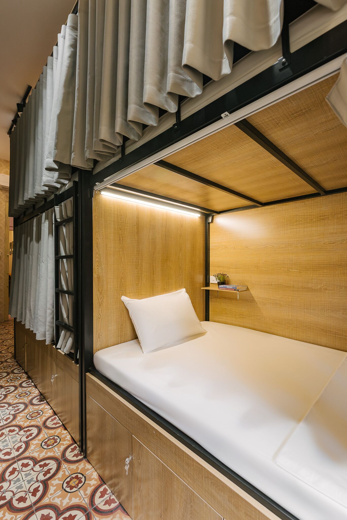 Una cama litera con cabecera de madera, un estante pequeño con libros y una planta, y baldosas de suelo con diseños.