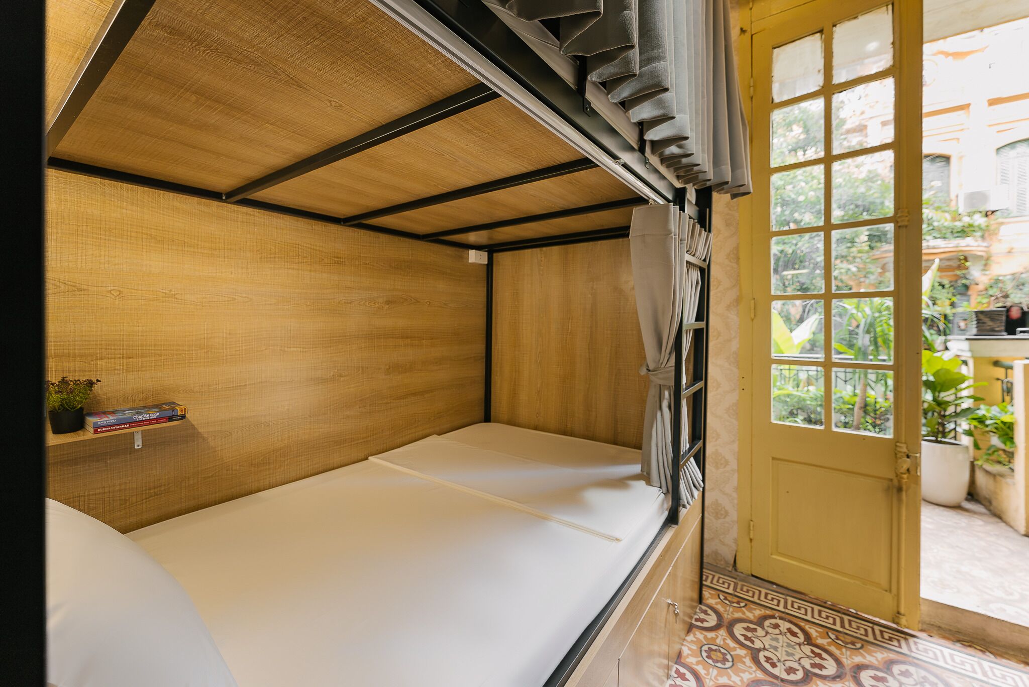 A compact room with a bed, a small shelf with books, and a door leading to an outdoor area with plants.