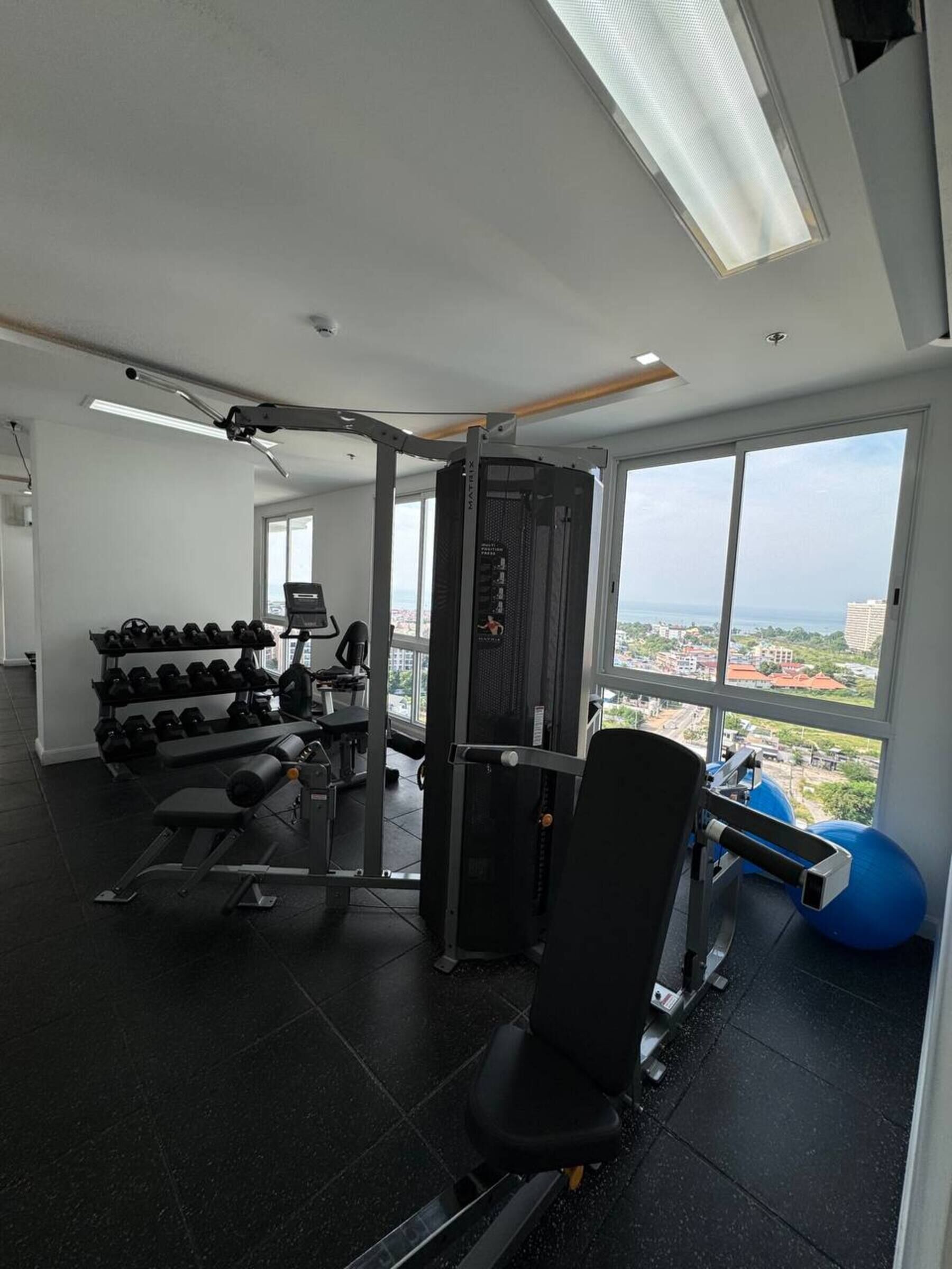 A gym with various exercise equipment, a rack of dumbbells, and large windows offering a city view.