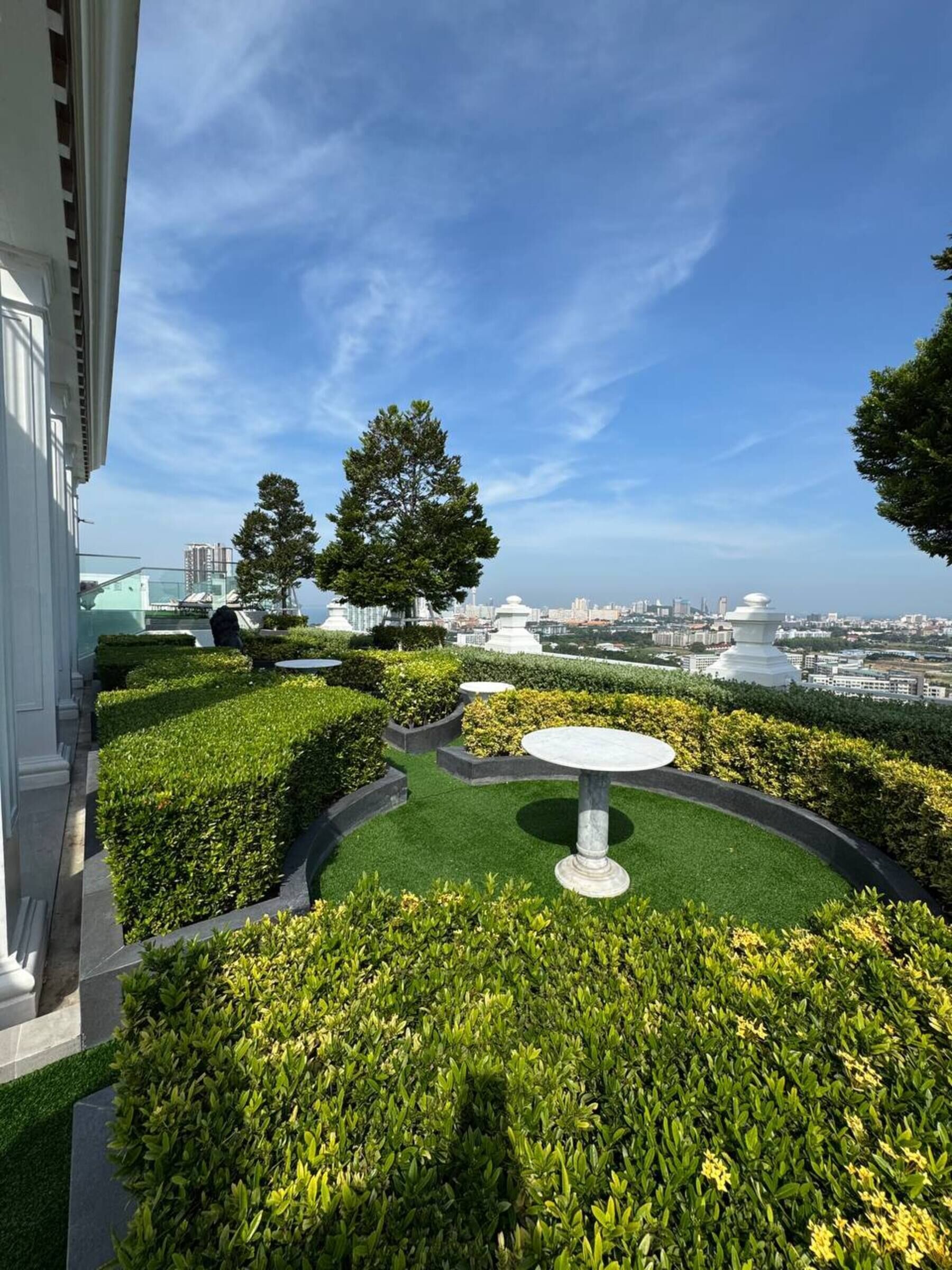 A rooftop garden with a central table, manicured hedges, and a view of the city skyline.