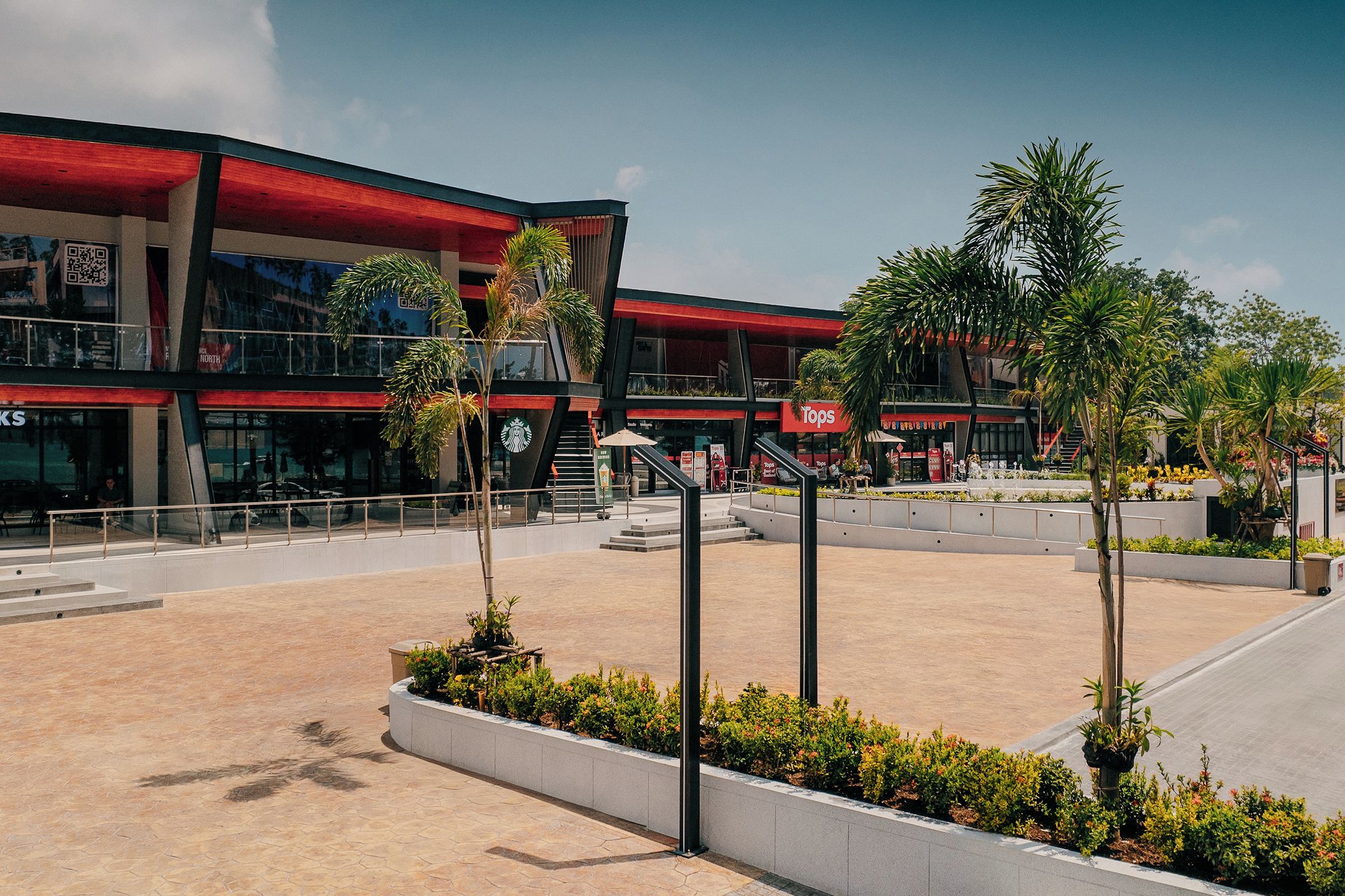 Un centre commercial moderne comprenant un Starbucks, un magasin Tops, et une place centrale ouverte agrémentée de palmiers et d’espaces aménagés pour s’asseoir.
