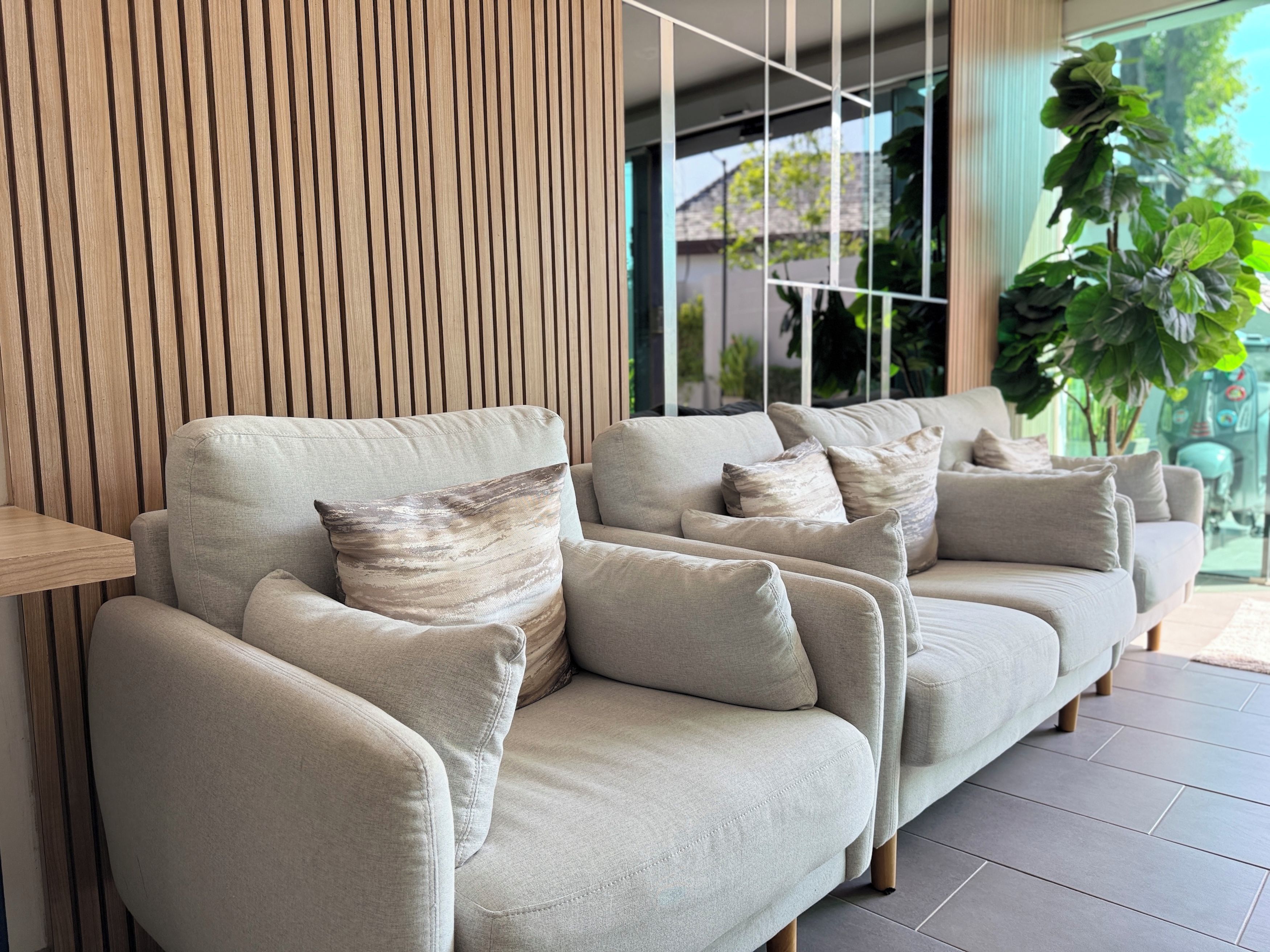A modern living area with a sofa set, a wooden slatted wall, and a view of greenery outside.