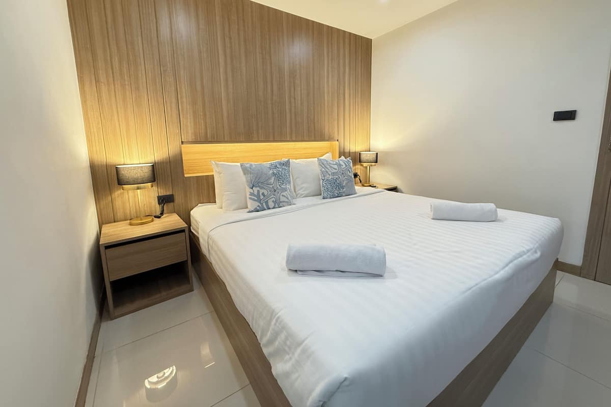 A modern hotel room with a wooden headboard, a neatly made bed with white linens, two bedside lamps, and a small wooden nightstand.