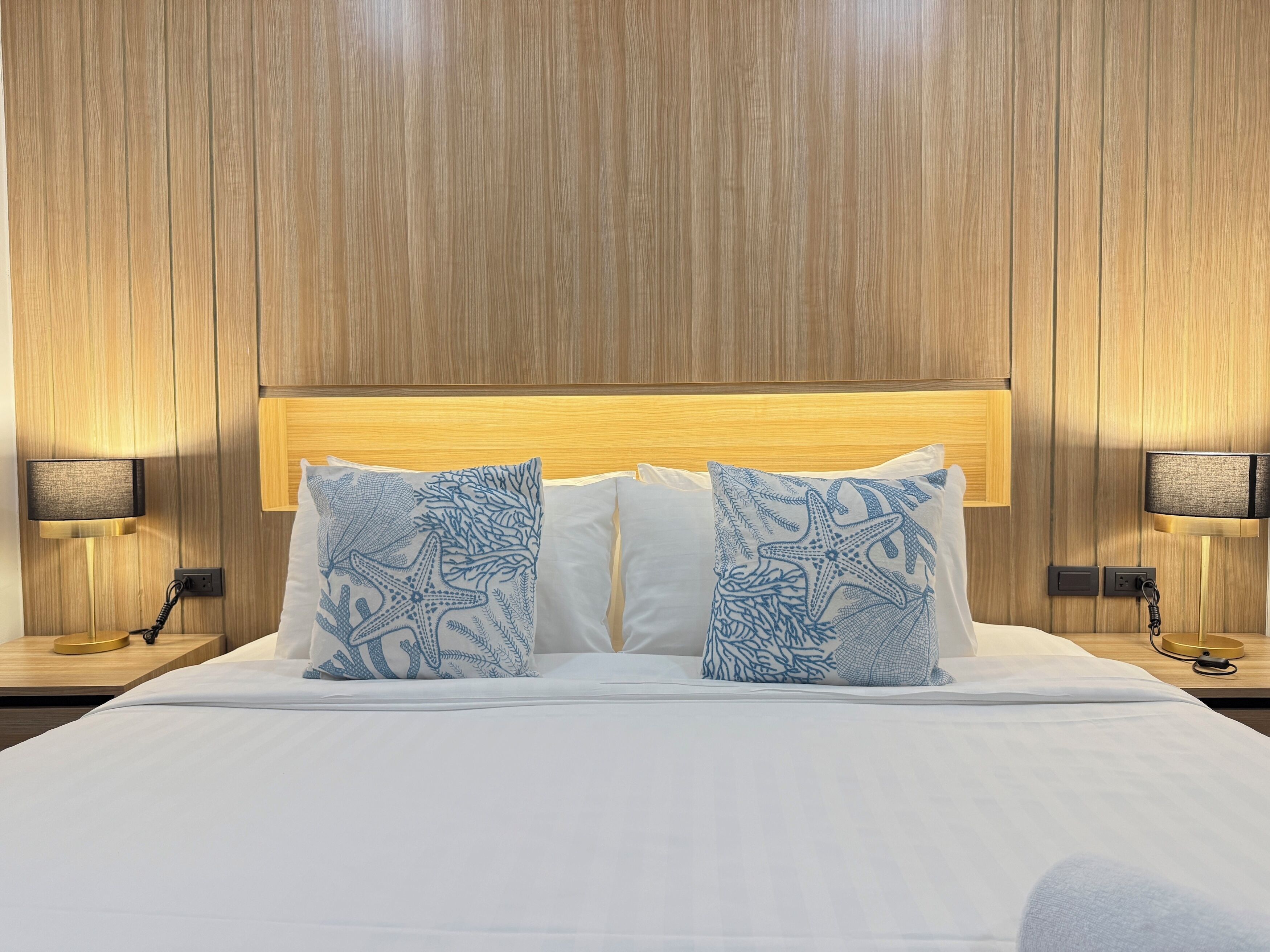 Une chambre d’hôtel avec une tête de lit en bois, du linge de lit blanc et deux coussins décoratifs à motifs bleus.