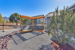 Une pergola en bois avec un banc, située dans un espace recouvert de gravier, entourée d’arbres et de maisons.