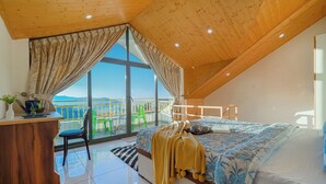 Een slaapkamer met een groot raam, een houten bureau, een bed met een bloemenprint en een balkon met uitzicht op de bergen.