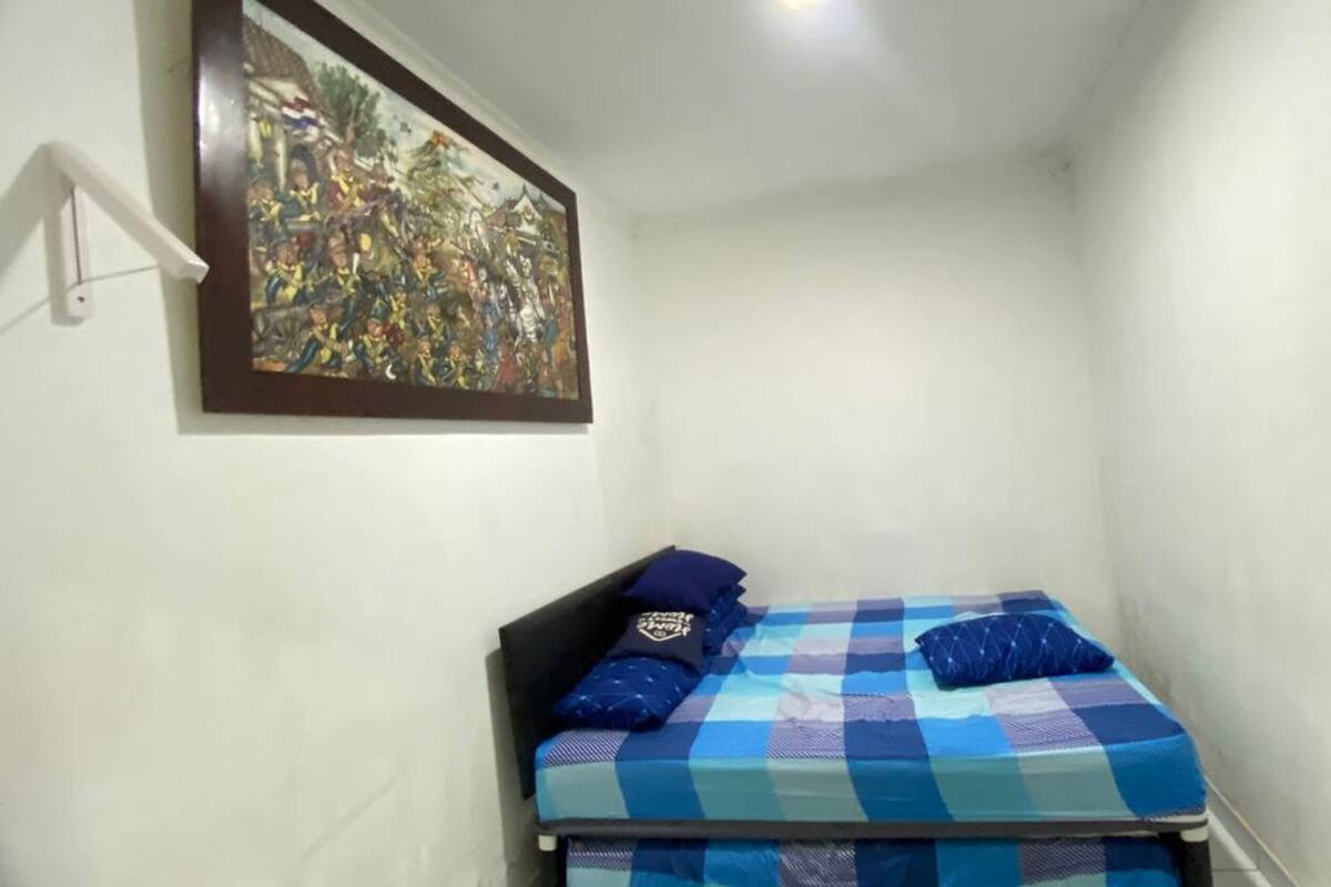 Ein kleiner, schmaler Raum mit einem Bett, das eine blau-weiß karierte Tagesdecke und zwei Kissen hat. Über dem Bett hängt ein gerahmtes Bild.
