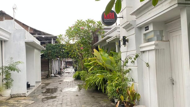 Une ruelle étroite bordée de bâtiments blancs, une enseigne indiquant « KARA HOMESTAY », et une végétation luxuriante le long du chemin.