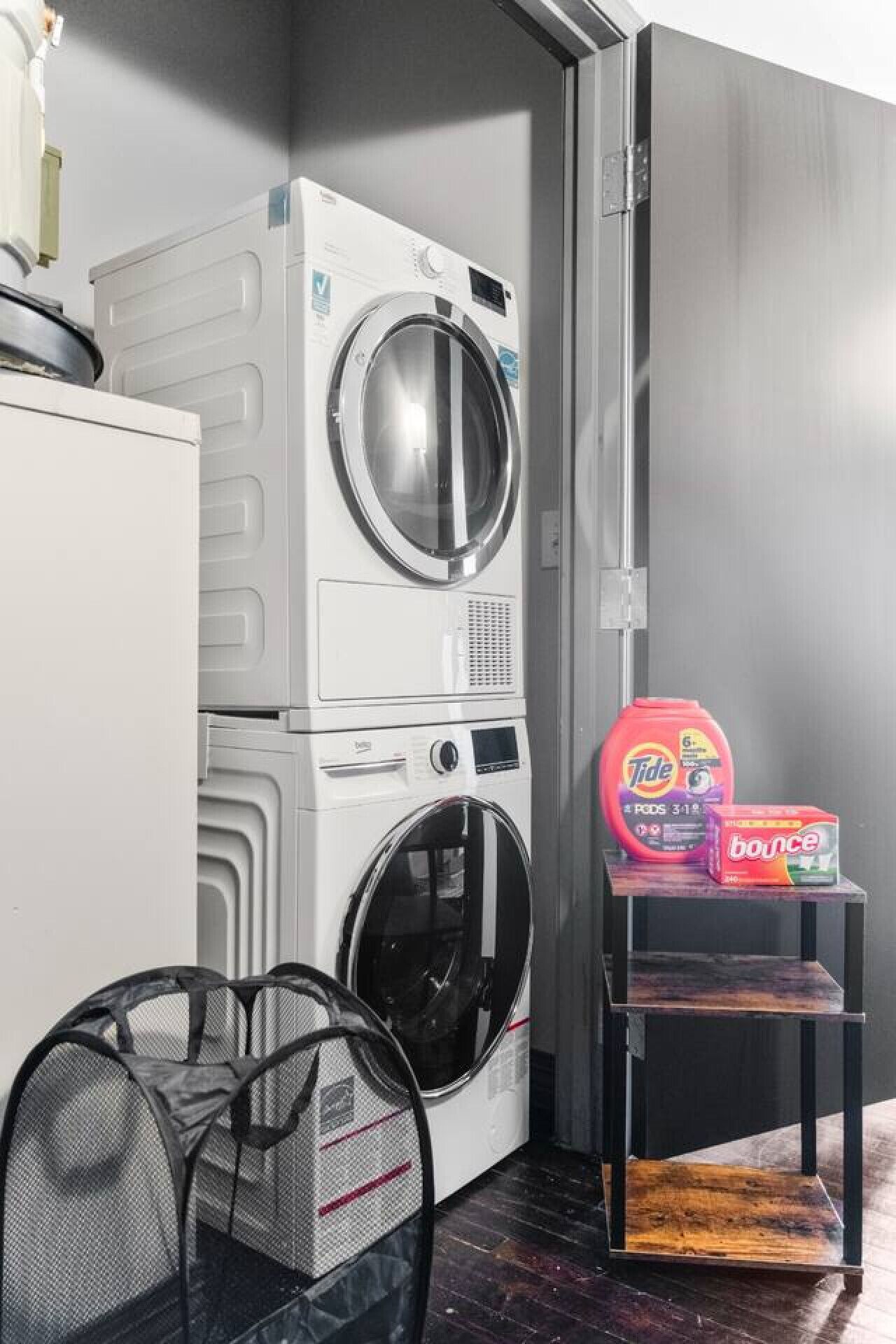 Un espace buanderie compact comprenant un lave-linge et un sèche-linge à chargement frontal, une étagère avec de la lessive et un panier à linge en maille.