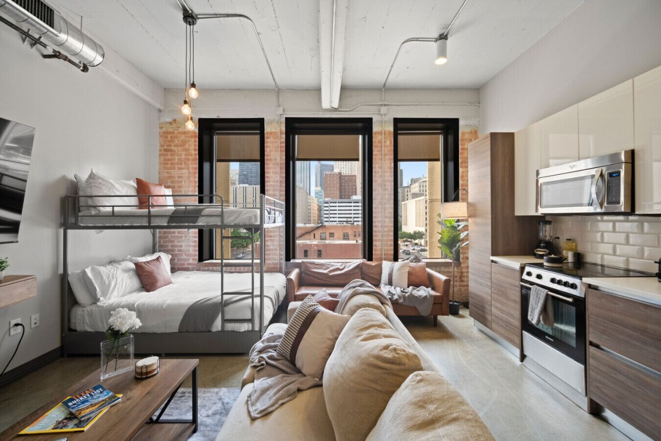 A compact studio apartment with a bunk bed, a sofa, a kitchen area with a microwave and stove, and large windows offering a city view.