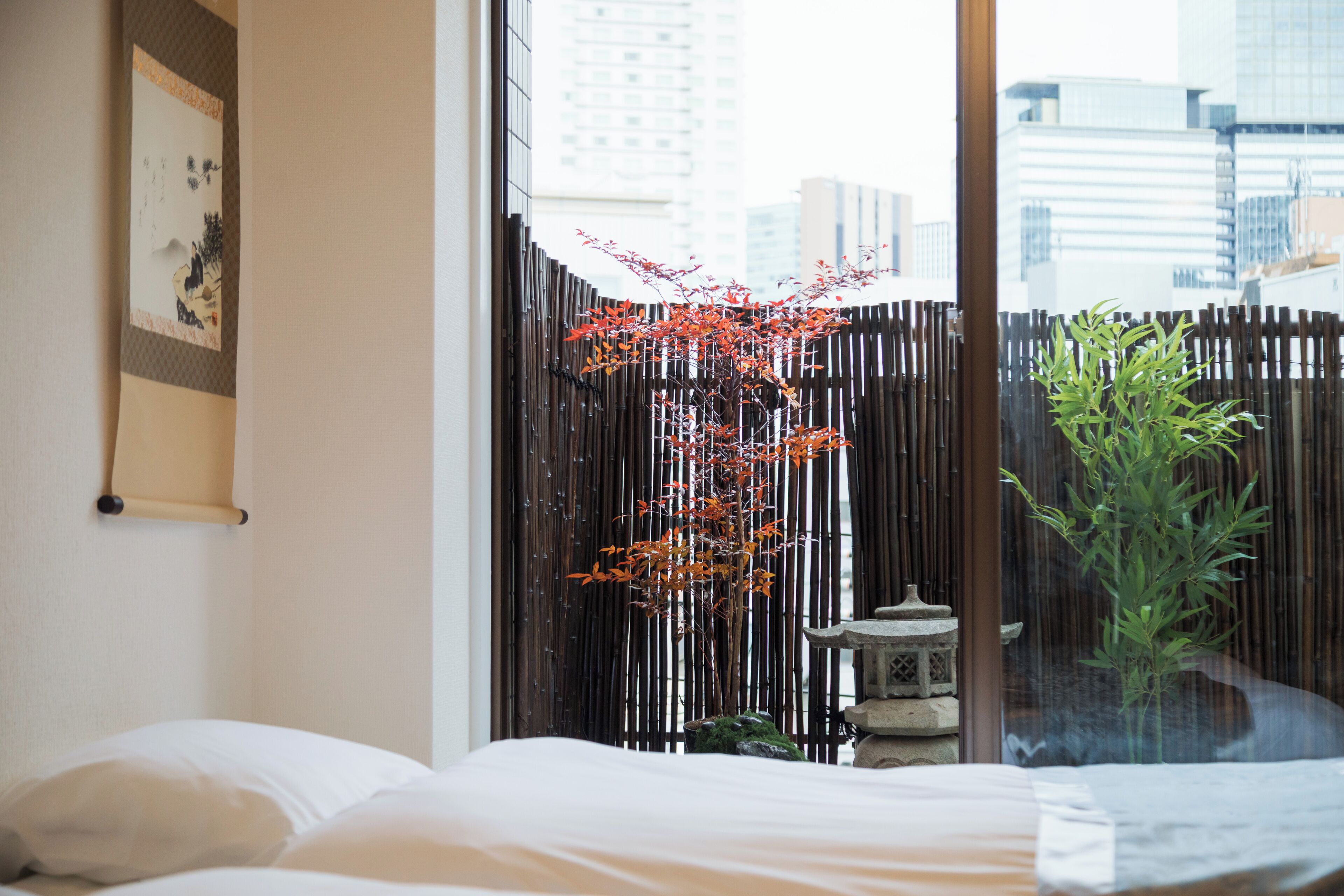 A hotel room with a bed, a window with a view of buildings, a bamboo fence, and a bonsai tree.