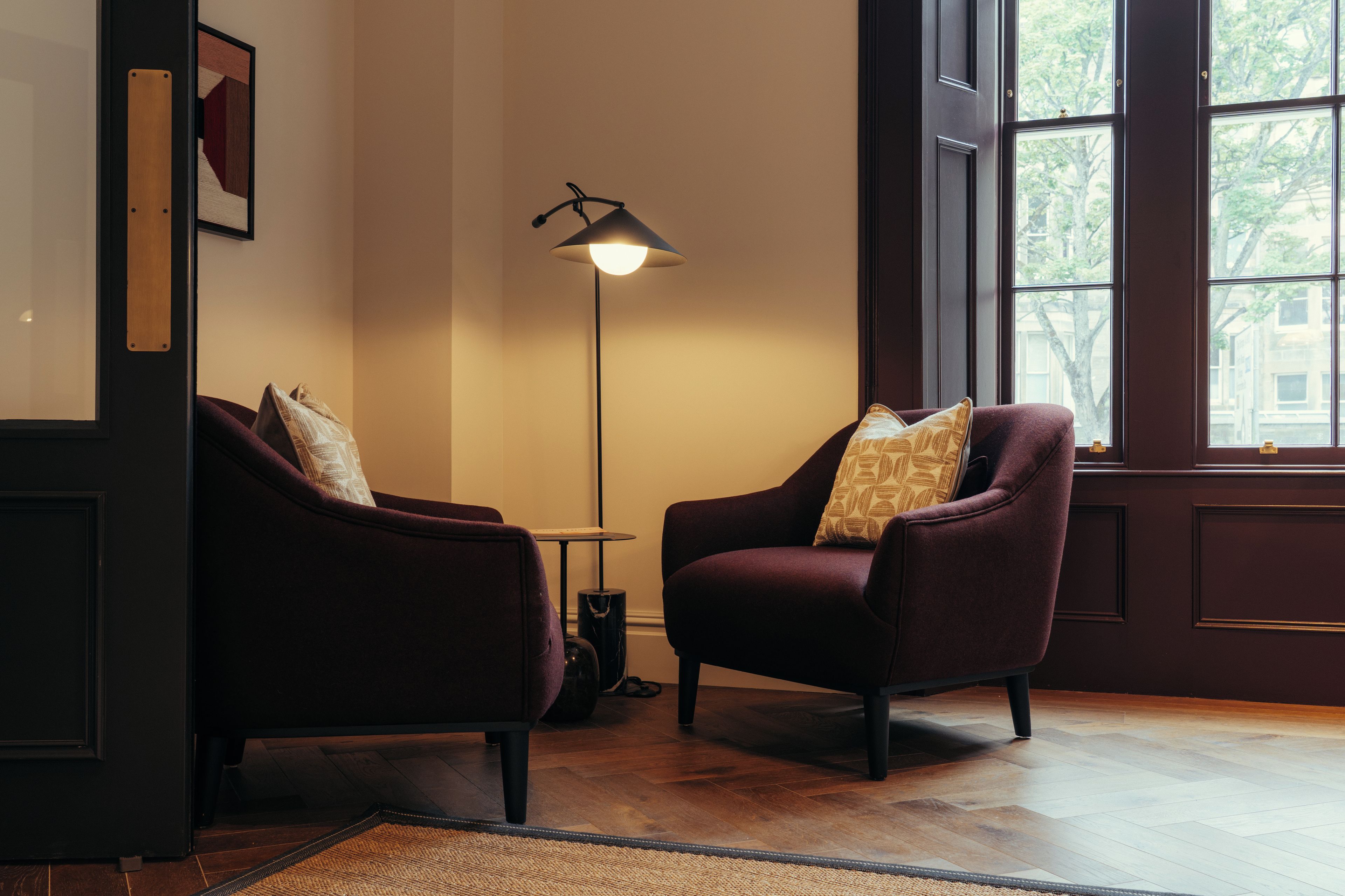 Una habitación con dos sillones, una lámpara de pie, una mesita y un ventanal con vistas a árboles.