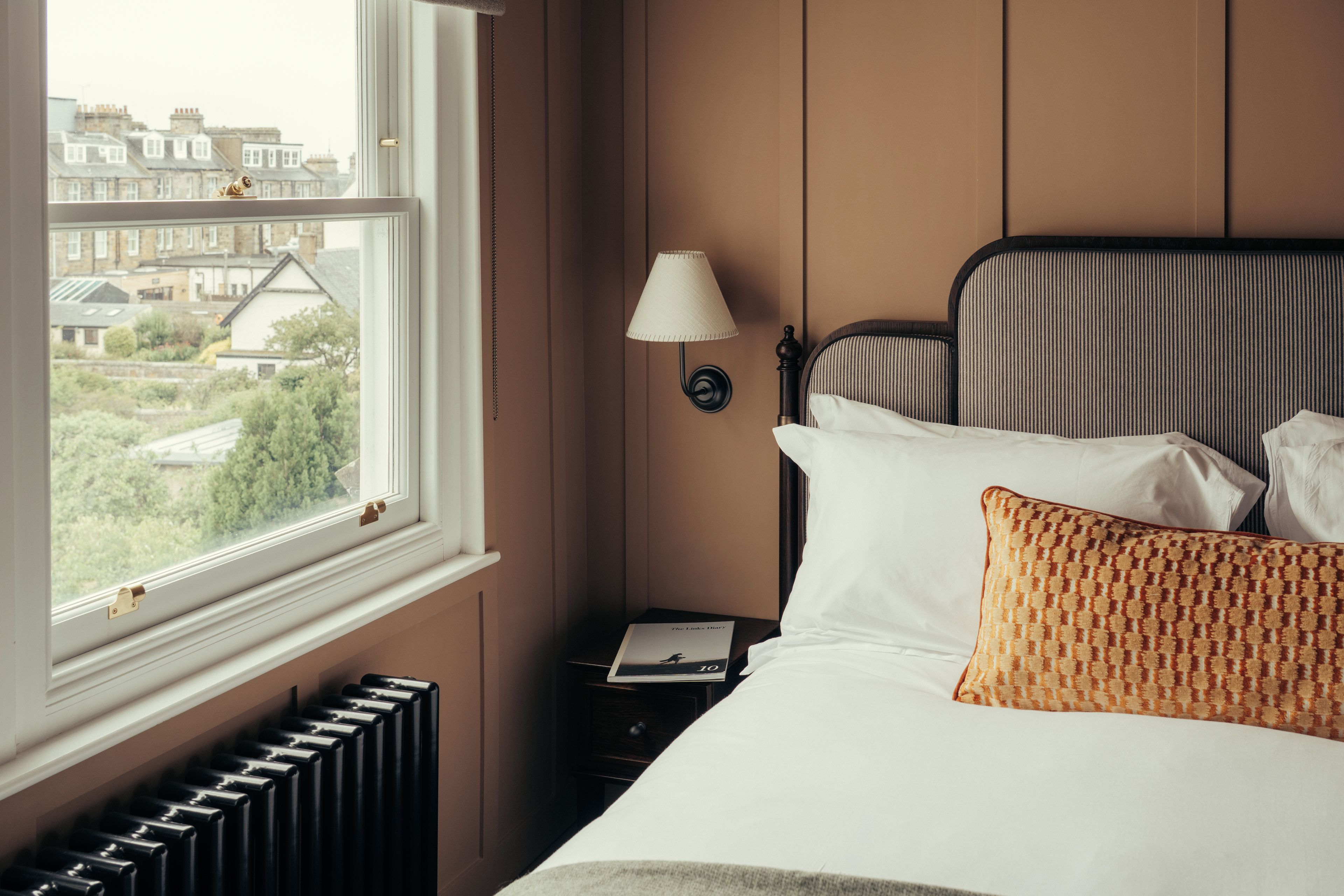 Ein Hotelzimmer mit Bett, Nachttisch, Lampe und Fenster mit Blick auf Gebäude.