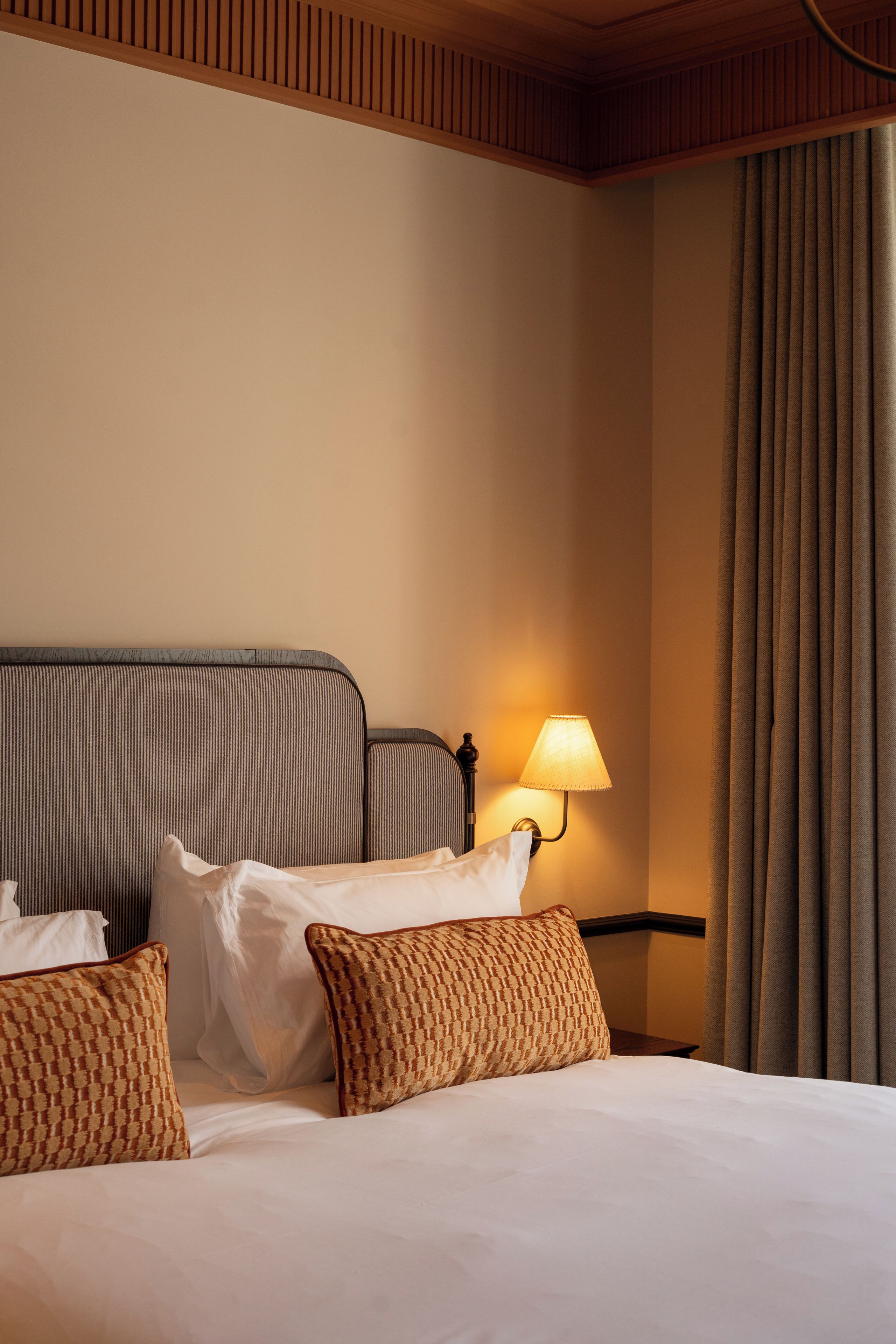 Een hotelkamer met een bed, kussens, een nachtkastje, een lamp en een raam met gordijnen.