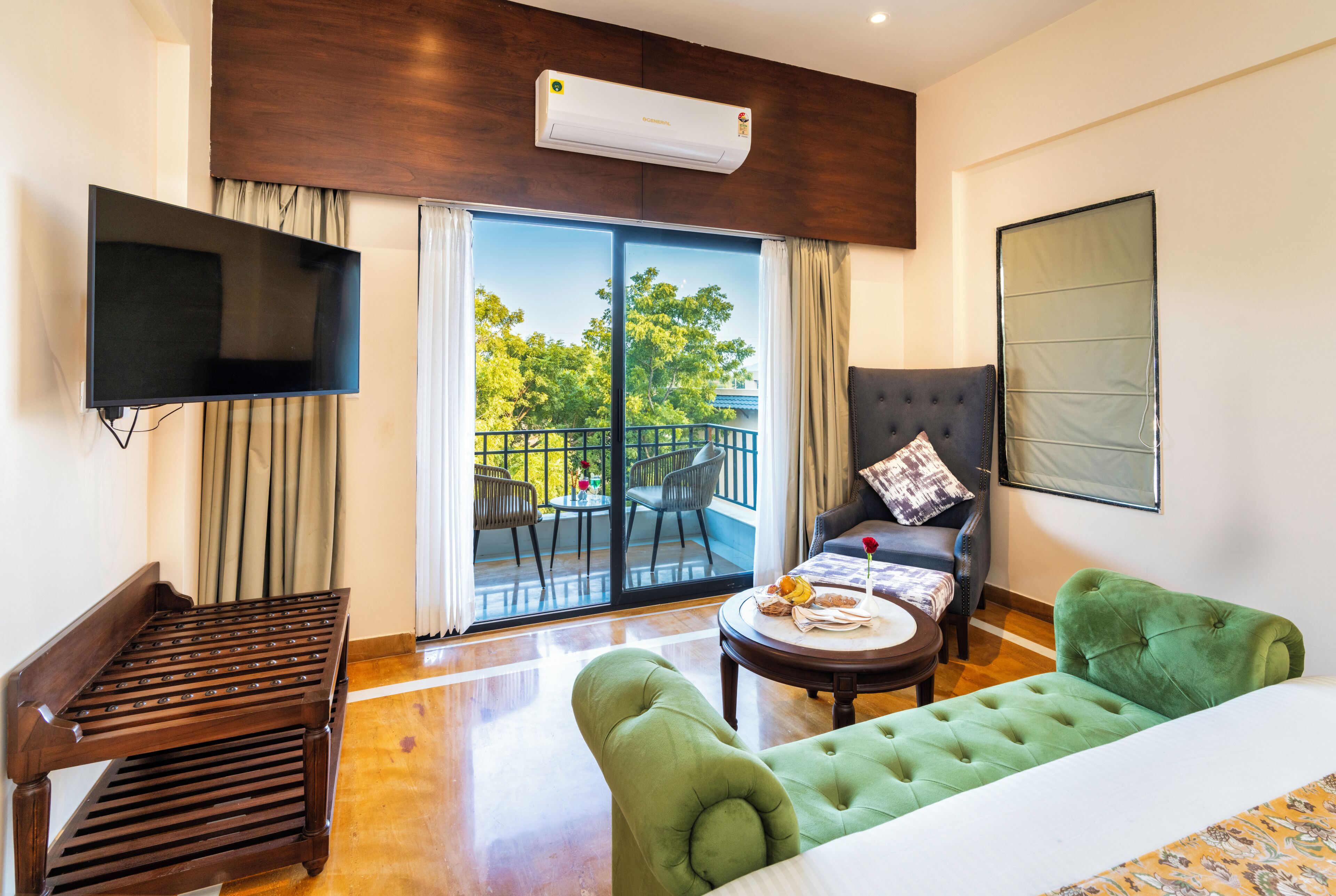 Habitación de hotel con televisor de pantalla plana, sofá, sillón verde, mesa de comedor y balcón con mobiliario exterior.