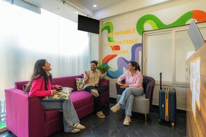 Un grupo de personas sentadas en un sofá rojo en una habitación con un ventanal grande, una pared con texto colorido y un ordenador portátil sobre una mesa.