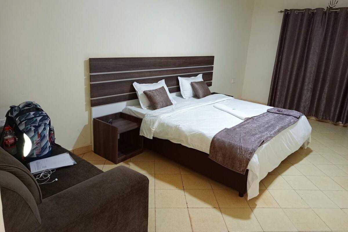 A neatly made bed with a headboard, a bedside table with a lamp, a chair, and a backpack on the floor.