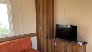 A compact room with a wooden cabinet, a television, a cushioned bench, and a small table. - Mobile home with terrace and heated pool in the heart of the seaside resort (Port-la-Nouvelle)