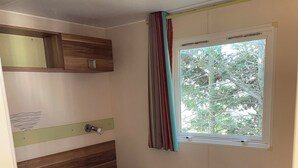 A single bed with a blue mattress, a wooden headboard, a window with curtains, and a wall-mounted shelf. - Mobile home with terrace and heated pool in the heart of the seaside resort (Port-la-Nouvelle)