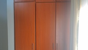 Ein hölzerner Kleiderschrank mit zwei Türen und zwei Schubladen, vor einer Wand mit einem Fenster zur rechten Seite.