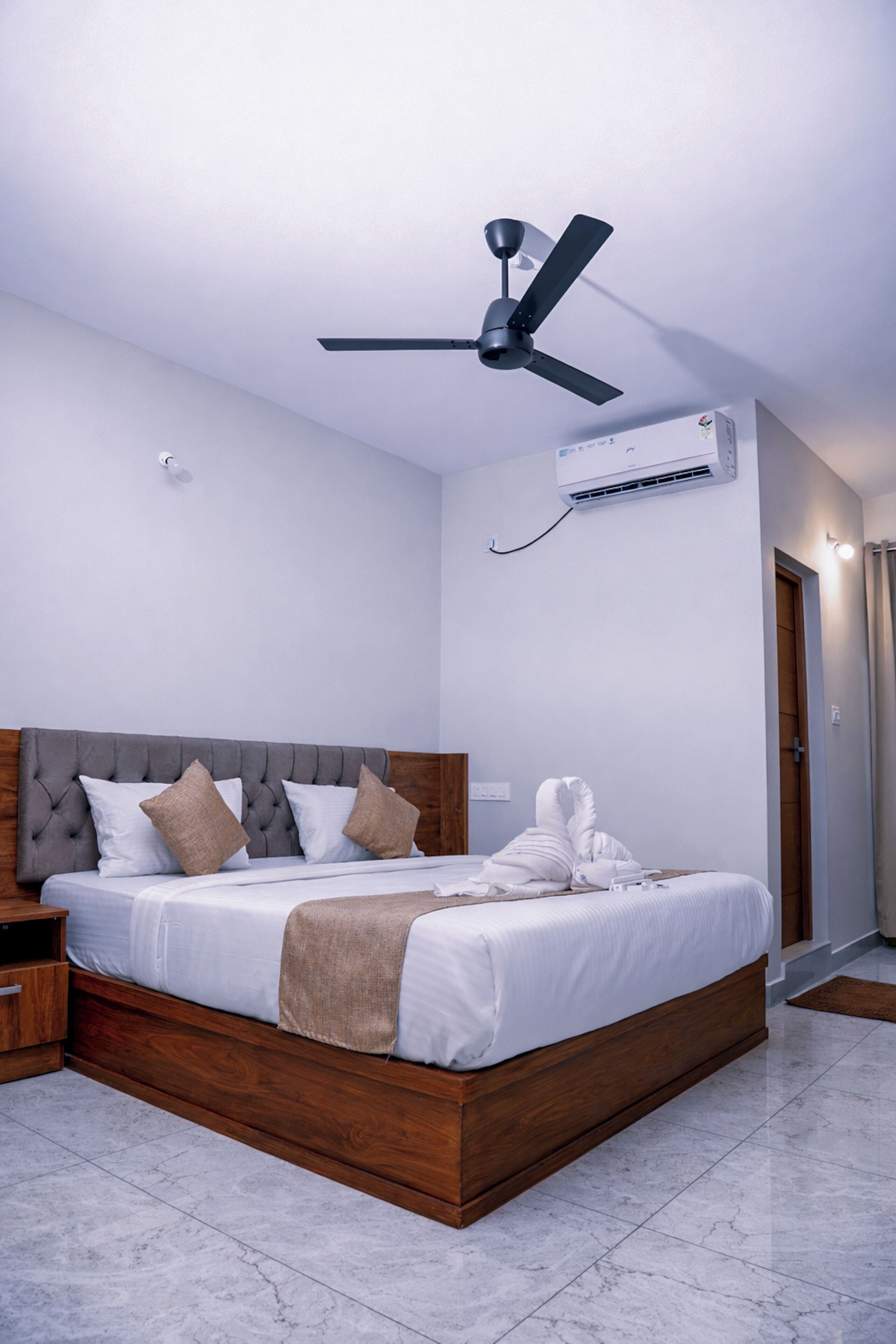 Une chambre d’hôtel moderne avec un cadre de lit en bois, un ventilateur de plafond, un appareil de climatisation et une table de chevet en bois.