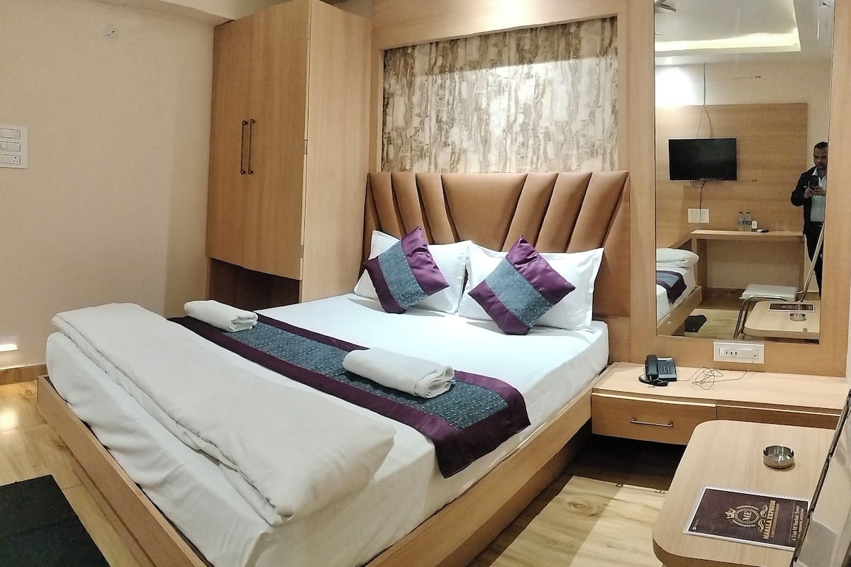 A modern hotel room with a large bed, wooden headboard, a bedside table, a mirror, and a small desk with a chair.
