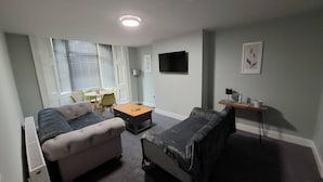 Un espace de vie compact comprenant un canapé, une petite table, un téléviseur fixé au mur et une fenêtre avec des stores.