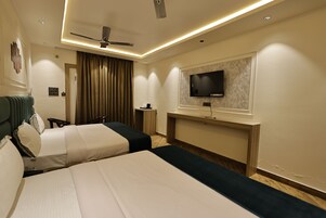 Habitación de hotel con dos camas, ventilador de techo, televisor de pantalla plana en la pared y una mesita.
