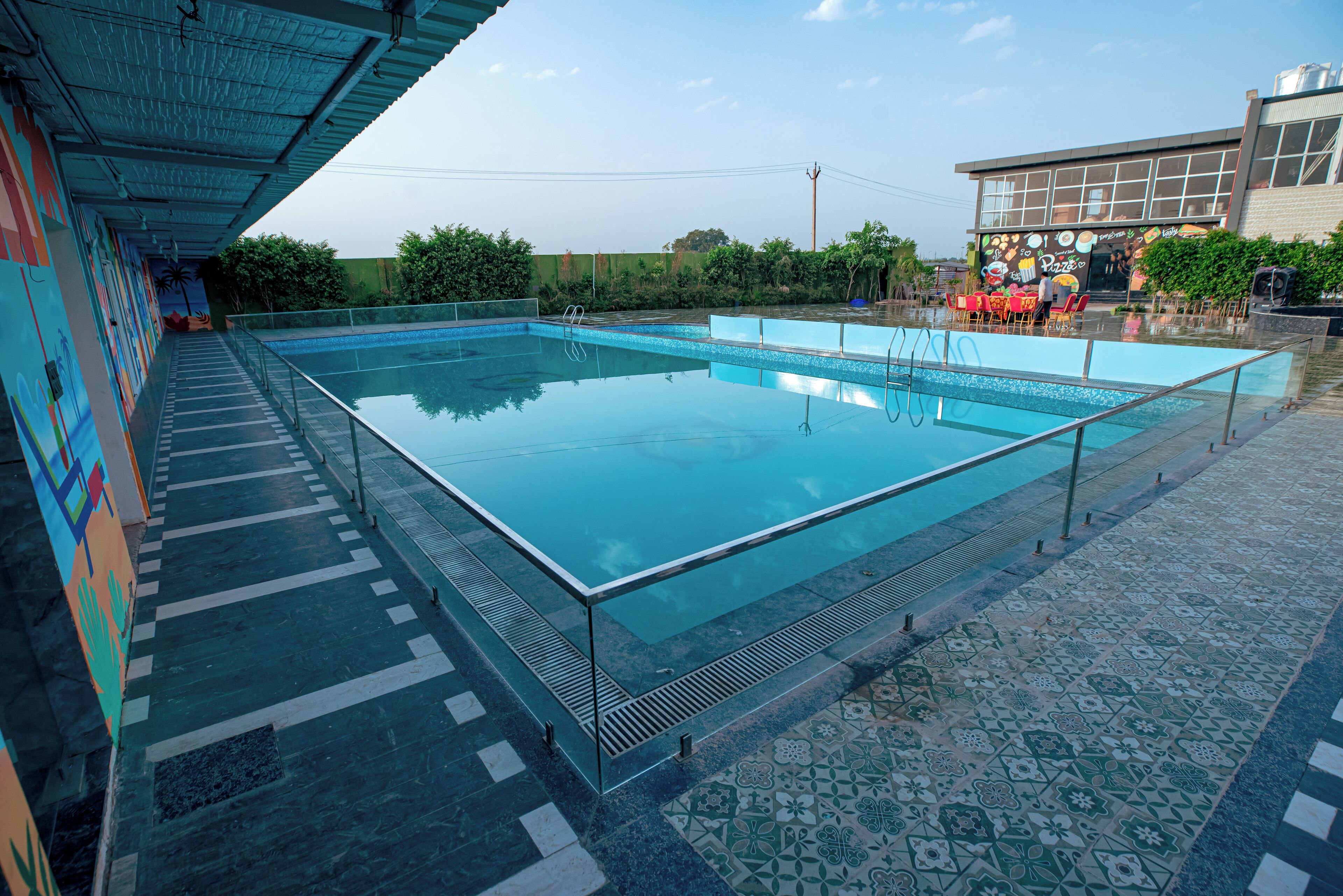 A swimming pool with a glass edge, surrounded by a tiled walkway and a covered area with a mural.