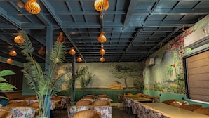 Een restaurantinterieur met hoge plafonds, hangende pendelverlichting, tafelkleden met bloemenprint en een vloer met patroon.