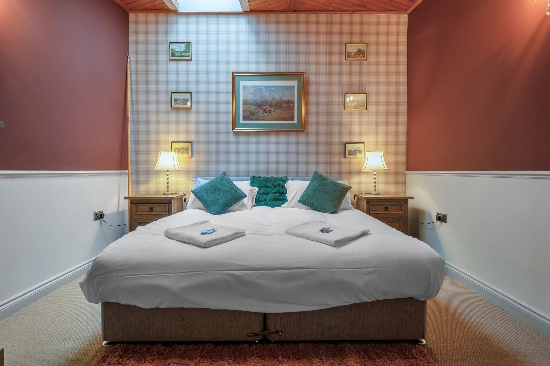 A neatly made bed with white linens, two green pillows, and a framed picture above it. There are two bedside tables with lamps, and the walls are adorned with a plaid pattern.