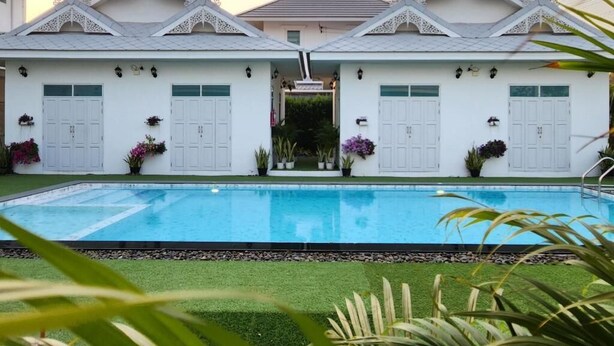 A poolside view of a building with white walls and decorative roof elements, a swimming pool, and potted plants. - Bianco House (Cha-am)