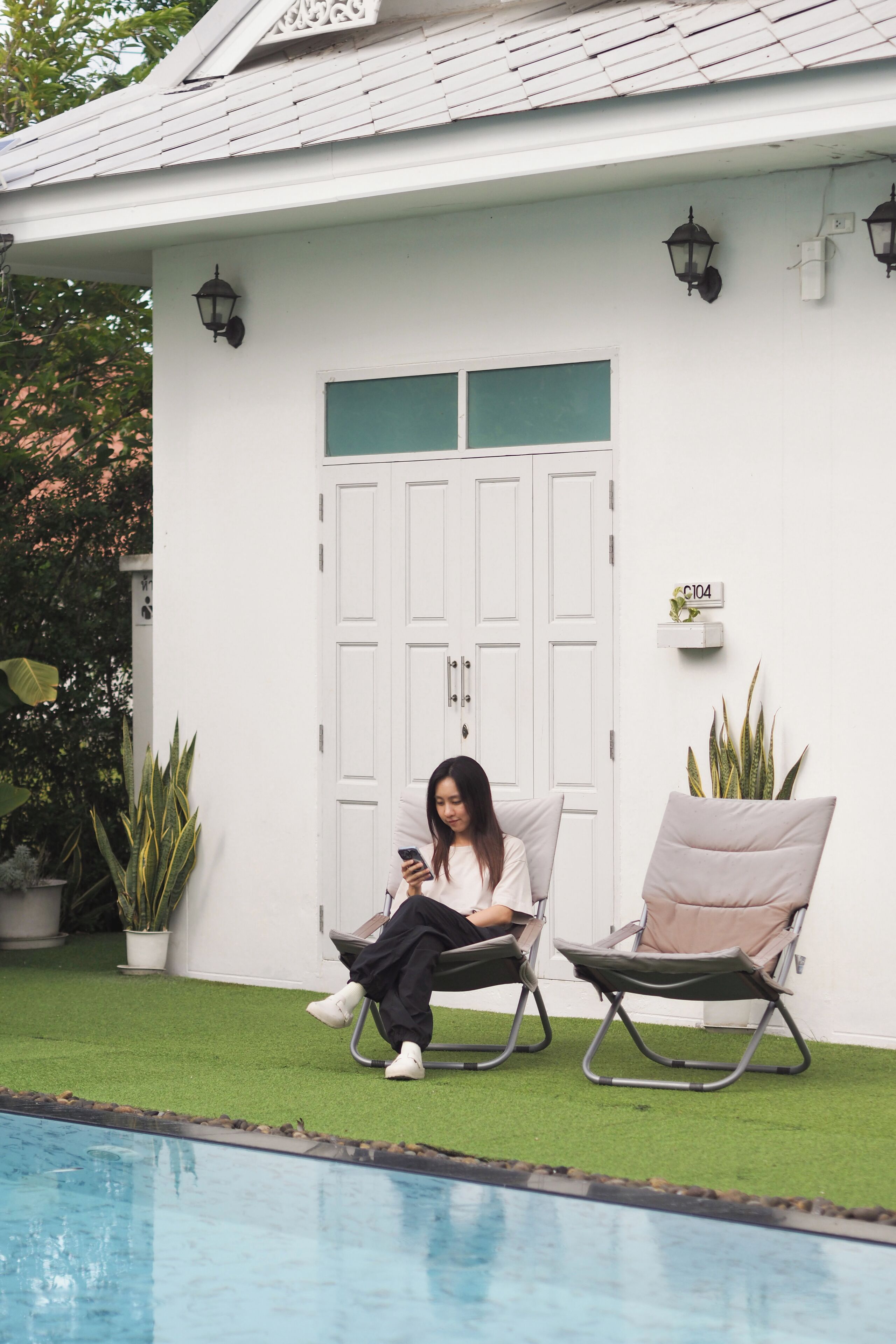 Una persona sentada en una silla de descanso junto a una piscina, con un edificio blanco al fondo y una puerta con un panel verde.