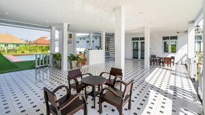 A spacious outdoor patio with a tiled floor, surrounded by white pillars, featuring a round table and chairs, and a view of a swimming pool and greenery in the background. - Bianco House (Cha-am)