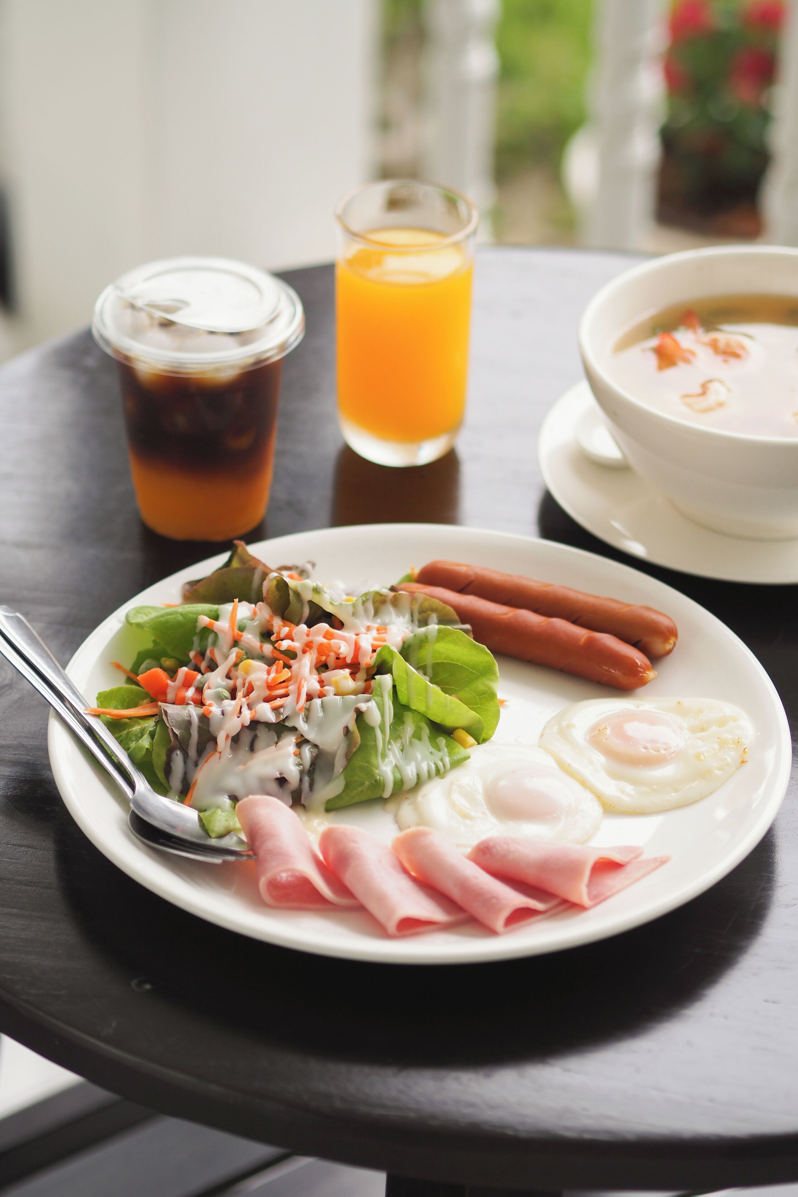 A plate with a salad, two sausages, a fried egg, and ham slices.