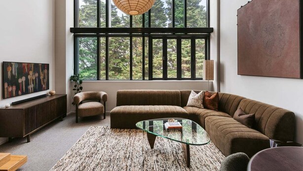 A modern living room with a large window, a brown sofa, a glass coffee table, and a painting on the wall. - Tree Tops Loft - Stylish, Private 2-Bedroom Retreat in Mount Maunganui (Mount Maunganui)