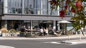 An outdoor café with tables and chairs, a bicycle, and a building with large windows in the background. - Tree Tops Loft - Stylish, Private 2-Bedroom Retreat in Mount Maunganui (Mount Maunganui)