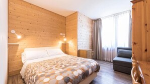 A room with wooden panel walls, a bed with a patterned bedspread, a nightstand, a dresser, a gray sofa, and a window with curtains. - Hotel Vescovi (Asiago)