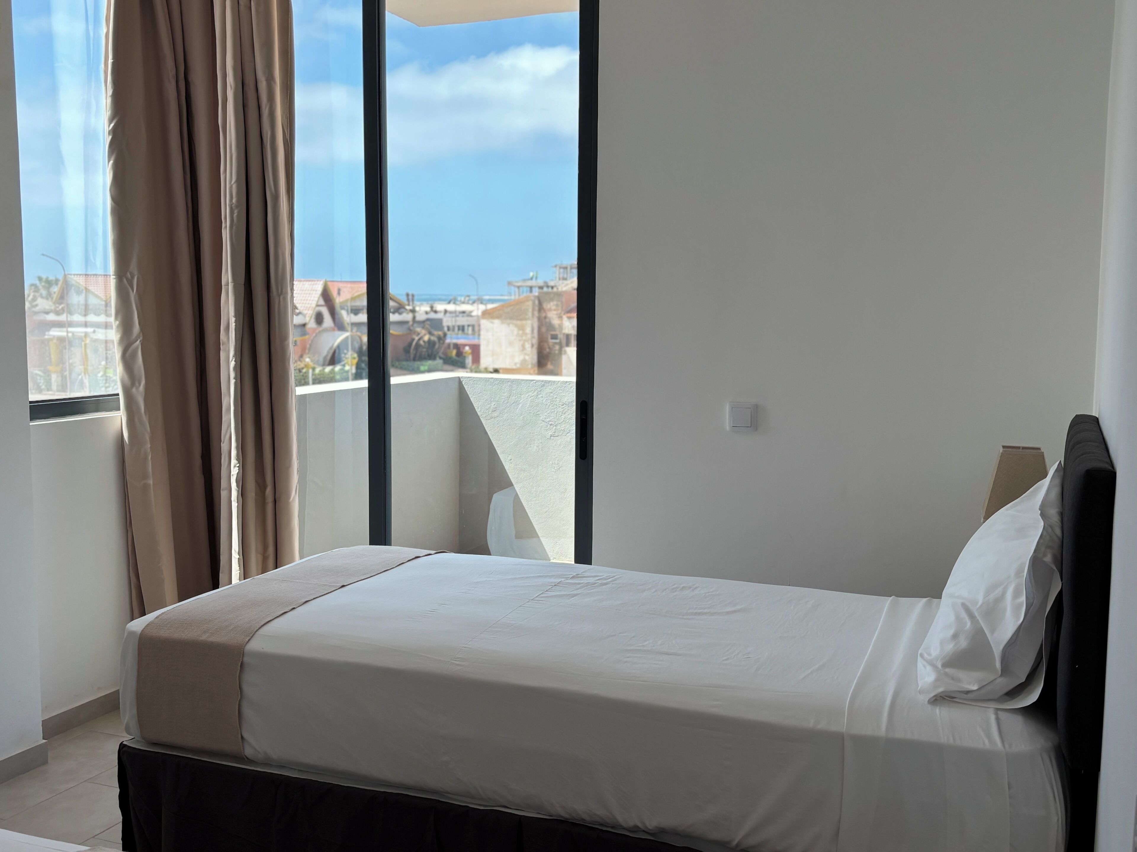 A hotel room with a bed, a window with curtains, and a view of buildings outside.