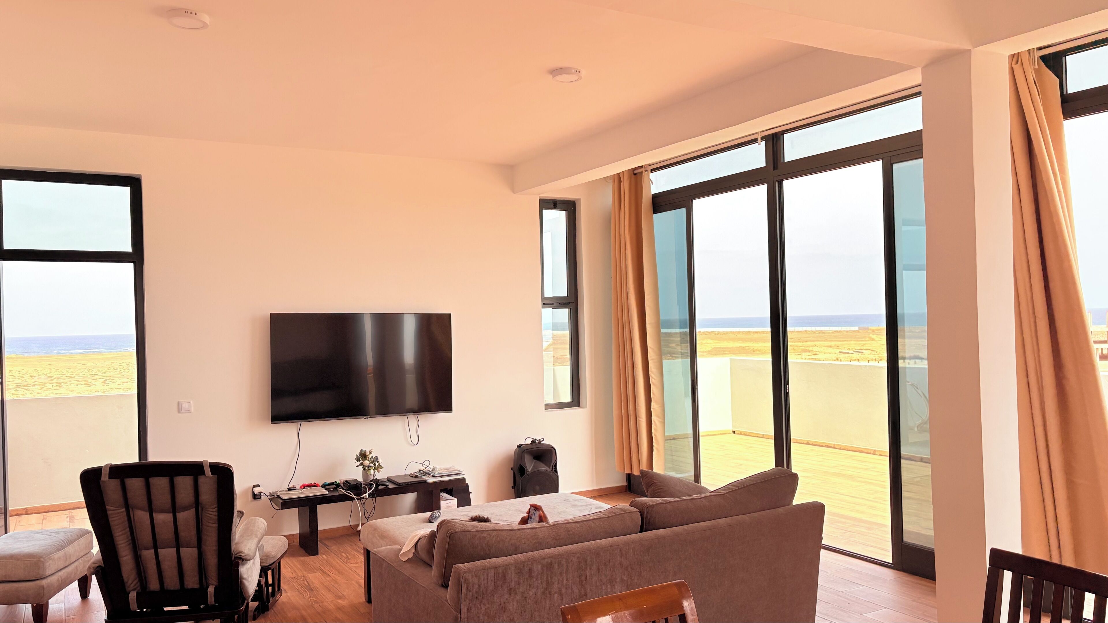 A living room with a flat-screen TV, a sofa, a chair, a small table, and a balcony with a view of the beach.