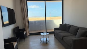 A modern living room with a large window, a flat-screen TV, a sofa, and a small table. - Joli Beach Apartments With Shared Pool (Sal)