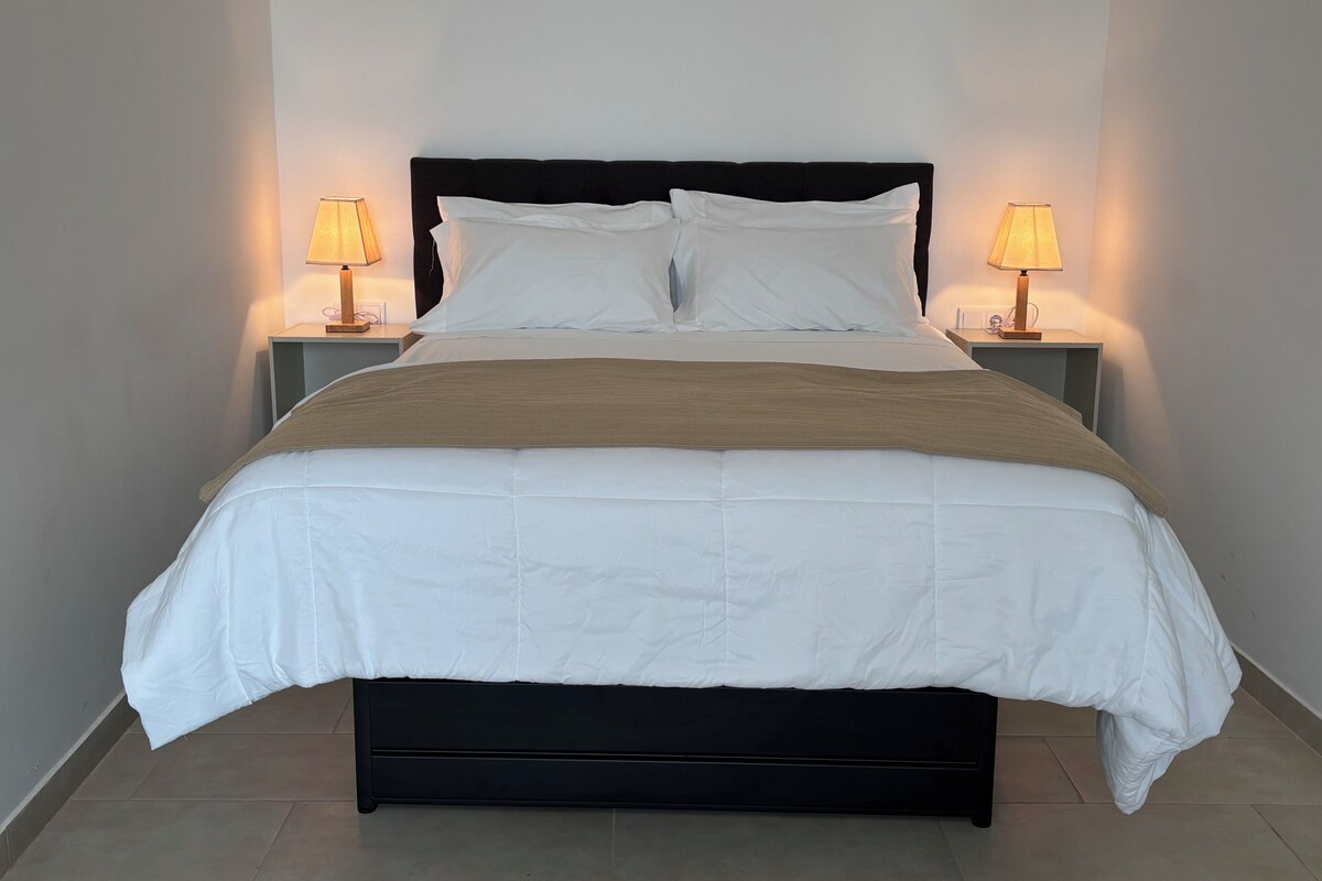 A neatly made bed with white bedding and a brown throw, flanked by two bedside tables with lamps.
