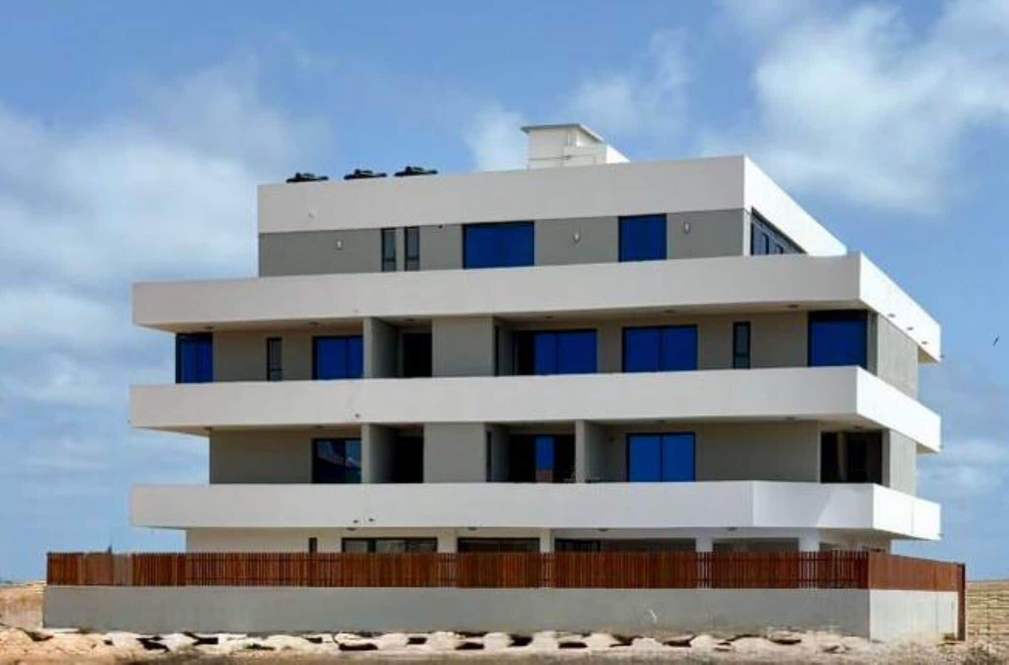 A multi-story building with a flat roof, a wooden fence at the base, and a clear sky in the background.