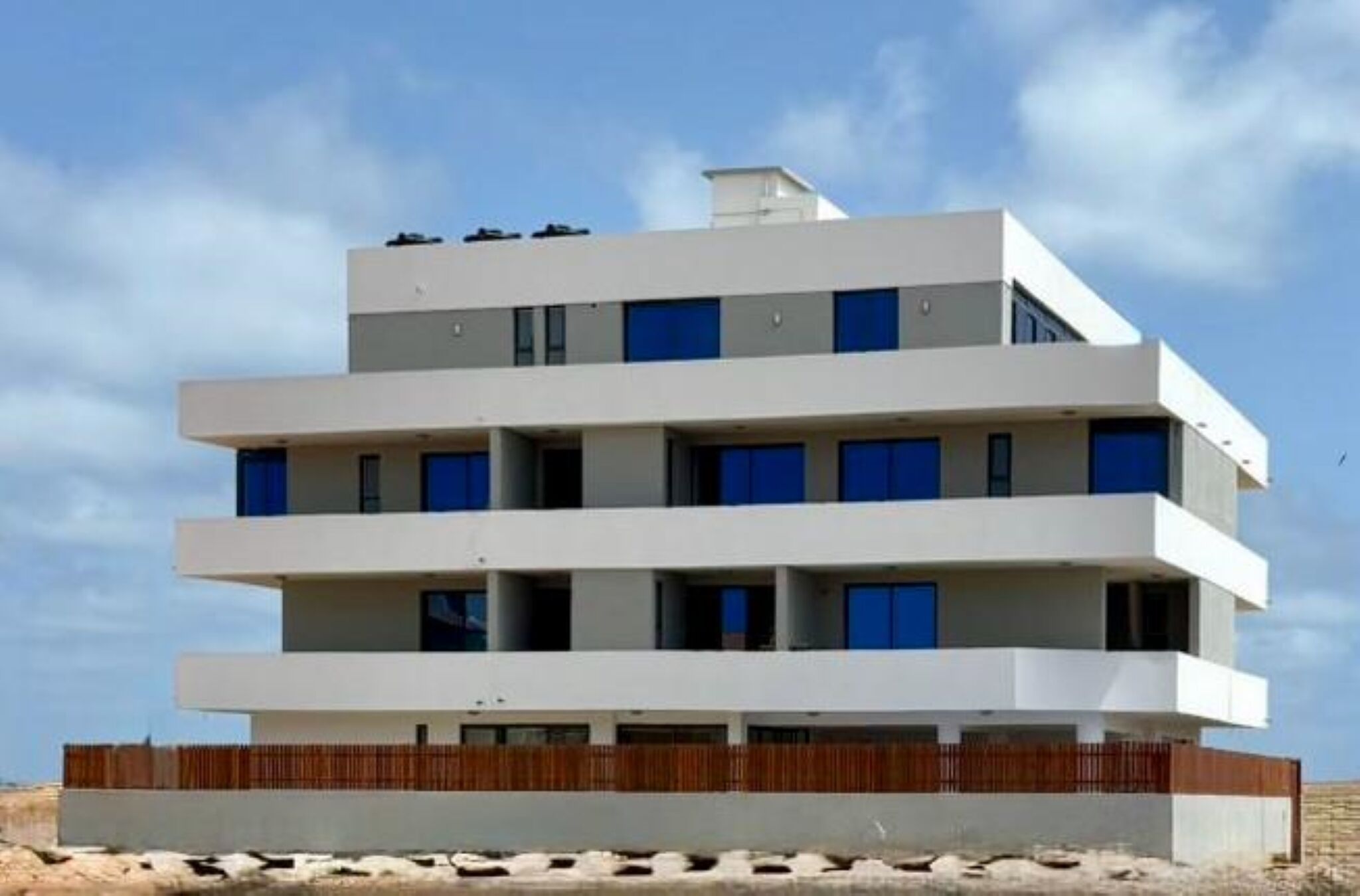 Un edifici de diverses plantes amb teulada plana, una tanca de fusta a la base i un cel blau al fons.