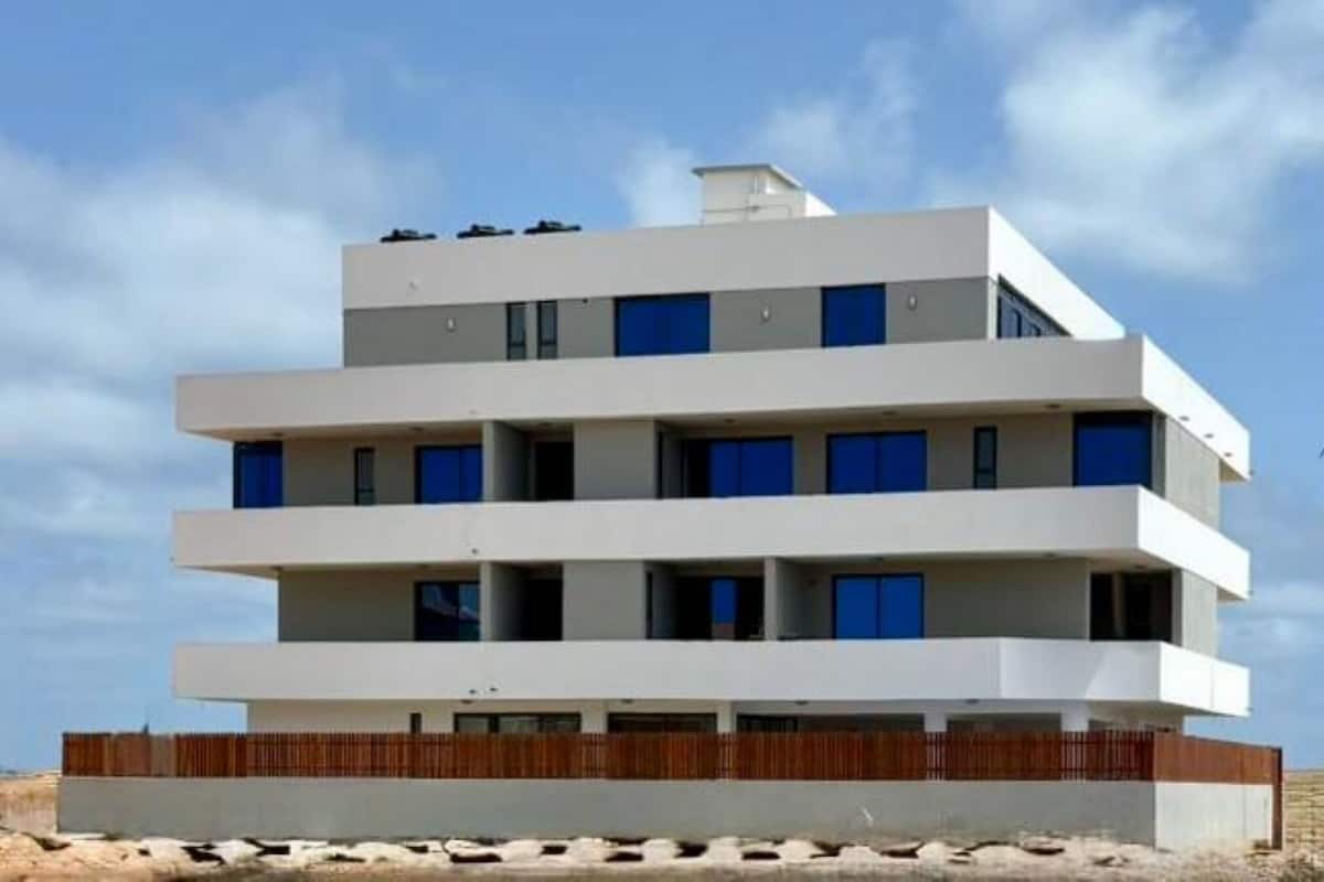 Un edifici de diverses plantes amb teulada plana, una tanca de fusta a la base i un cel blau al fons.